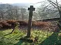 A tail, plain, short-armed cross on a plain base in a graveyard overlooking a trees and a green valley