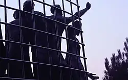 Close-up of sculpted human figures reaching through the fence
