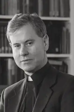 A black-and-white image of a young priest with short cropped hair in front of a bookshelf