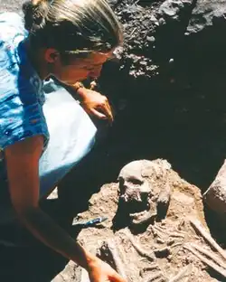 A young woman on an archaeological site, reaching down into a grave, where a human skeleton can be seen. She has her hair tied up behind her head and wears a patterned, short-sleeved shirt.