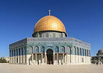 An octagonal, multi-colored building, the upper trim of which is inscribed in Arabic, topped by a golden-plated dome