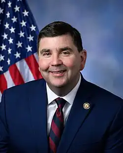 Mannion smiling in front of the U.S. flag, wearing a blue suit with his House member pin, a white shirt, and a red and blue striped tie.