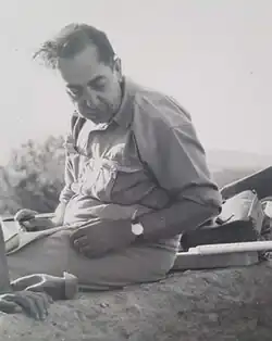 Papadimitriou, in middle age with coiffed black hair, sits on the side of an excavation trench. He wears shirt sleeves, pants and a wristwatch.