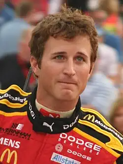 A man in his late twenties wearing black sunglasses with a head full of hair and a red jacket with sponsors' logos.
