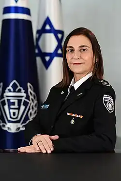 Katy Perry, a uniformed Israeli police officer, sits at a table with her hands folded, posing in front of an Israeli flag and the flag of the Israel Prison Service.