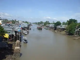 Brown-coloured waterway, with many small boats and elevated wooden and thatched houses visible. Foliage is also in abundance.