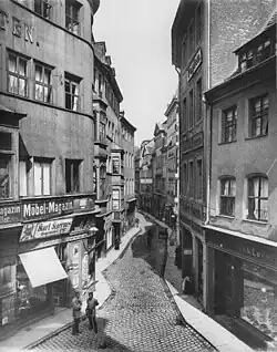 Black and white image of a narrow cobblestone street with stores alongside, two policemen are convened in the foreground
