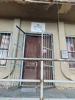 The entrance to a building, with a worn brown double door behind an open, grey metal security gate. A white sign with Georgian text and logos is mounted on the beige wall above the door.