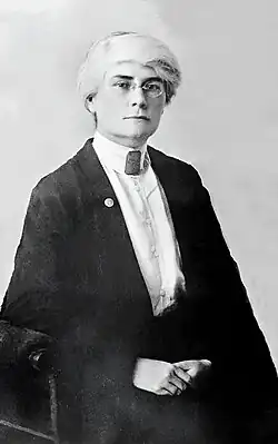 A white woman with white hair and dark eyebrows, wearing eyeglasses, a high-collared white blouse with many fabric-covered buttons down the front, and a dark jacket
