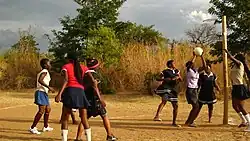Malawian young women playing netball on a dirt court. One player is preparing to shoot for goal, while other players in and around the shooting circle look on. Bibs are not worn by any of the players.