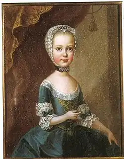 A young, blonde girl in a greenish-blue dress and a white cap. Her dress is embellished with white lace, greenish-blue bows and golden embroidery.