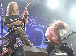 A color photograph of two members of the group Children of Bodom standing on a stage with guitars, drums are visible in the background. Both electric guitarists have "flying V" style guitars and they have long hair.