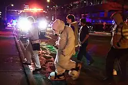 Nighttime photo of five people walking across a street. One is wearing a white fur suit and another a gas mask and high-visibility jacket. A fire engine is parked in the background with its lights on.
