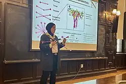 Mona Minkara stands in front of a scientific slide at Yale, holding a 3D printed model of surfactant protein D. The projected image shows structural domains and glycan-binding regions of the protein.
