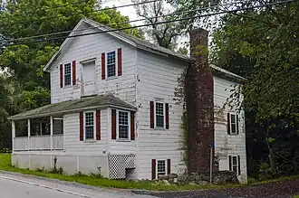 A white wooden house with clapboard siding and a front-gabled roof, seen at a three-quarter angle from across a road in front of it. Its windows are in an irregular pattern, with red wooden shutters and double-hung 12-over-12 sash windows. A porch comes off to its left to wrap around the far side of the house, and there is a door between the two windows on the second story facing the road. On the side a large brick chimney rises to the roof.