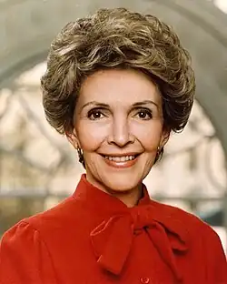 A headshot of Nancy Reagan, a smiling middle-aged woman with short hair dressed in red, facing the camera with a large arched window in the background.