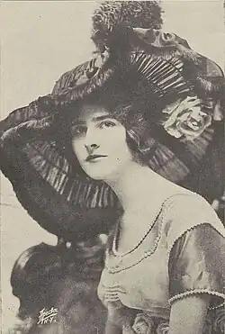 A young white woman wearing a large elaborate dark hat, and a gown trimmed with pearls