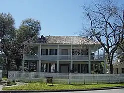 A gambrel-roofed house with large windows, wraparound balcony and first floor porch with Grecian columns. Sashed windows are framed by shutters.