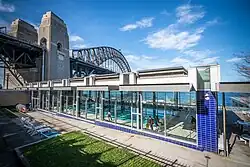 An indoor pool is seen through the windows of a glass building. In the background is the Sydney Harbour Bridge.