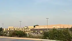 Campus parking lot with bushes and wide one-story building in the distance