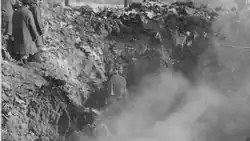 Men stand at the edge of a deep crater in the ground with billowing smoke coming from it