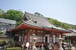 Wooden building with raised floor, white walls, red beams, a hip-and-gable roof and a canopy over the staircase.