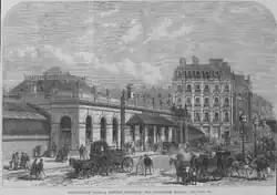 A busy street scene features a single-storey building with semi-circular arched windows and urns along the cornice. The pitched roof has ironwork details. A glazed awning shelters the entrance.