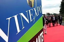 Close-up image of blue, green, and brown signage with "Vinexpo" in white lettering, with people walking on a red carpet visible on the right background