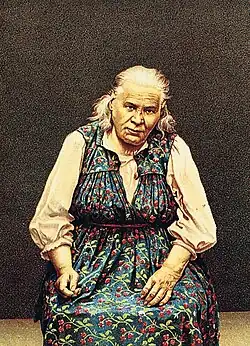 An old seated woman wearin a blue dress with red berries pattern and a pink blouse.