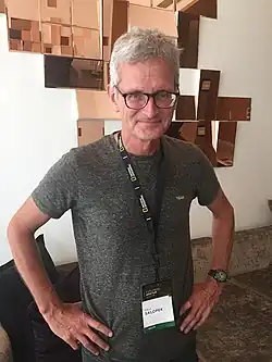 Paul Salopek wearing a gray t-shirt and a National Geographic conference lanyard with name, standing with arms akimbo, smiling directly at camera