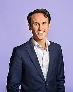 A smiling Caucasian man with dark hair in a suit in front of a purple background