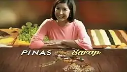 An image of Kara David sitting, with her hands on a wood table. Food ingredients are seen in the back. The show title is displayed on the image, below David's hands.