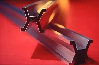 Two grey metal bars, one on top of the other, laying on a red table. The cross section of the bars is like an X with a central surface in the middle and with the tops and bottoms of the X flattened.