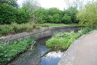 In contrast, a channelized part of the River Ravensbourne, with deliberate gravel shoals and plantings in its bed to make the flow meander and slow it down. This is cheaper than demolishing the concrete (see images below). The adjacent park also acts as a floodplain.[42][43]