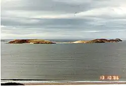 Two green sun-lit islets connected by a sandy tombolo sit offshore from a beach. The skies are grey and the sea all but flat calm.