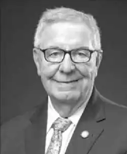 A monochrome portrait photo of a man with short grey hair, wearing a suit coat and tie; he is looking and looking straight into the camera with a happy expression.