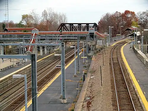 A railway station complex seen from a bridge. At left is a three-track line at ground level; at right is a single-track line rising on an embankment. Both lines have asphalt platforms. In the background, a footbridge and railway bridge over the ground-level line are visible.