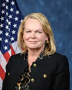 Official House portrait of McClain Delaney smiling in front of the U.S. flag, wearing a black blazer jacket and a gold and black shirt.