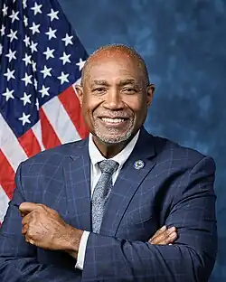 Official House portrait of Conaway smiling with his arms crossed in front of the U.S. flag, wearing a checkered blue jacket with an Air Force lapel pin, white shirt, and floral blue tie.