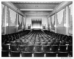 Black-and-white photo of the interior of the Revue Theatre, taken from rear of room facing stage with ornate curtains, art deco light fixtures, and narrow wooden chairs in rows with two aisles.