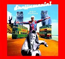 A photo of Hitchcock with his arms extended, standing between two buses, with a child seated in front of him and comically large cats behind him on a beach