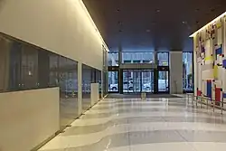 View ot the lobby, facing south toward 50th Street, along the passageway on the eastern side of the elevator core. There are white and gray pavers on the floor. To the right is a wall with a multicolored art piece with geometric spaces.