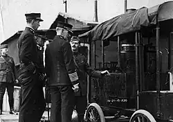 Four uniformed soldiers in front of a car, a uniformed soldier and a building with chimneys in the background. Black and white photograph