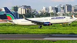 A US-Bangla Airlines Boeing 737 aircraft with tail number S2-AJA is taking off from a runway. In the background, a cityscape of modern high-rise buildings is visible. The runway is surrounded by green grass.