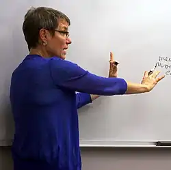 A photo of Sally Haslanger delivering a talk or lecture at a whiteboard.