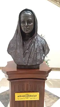 alt=statue of Savitribai Phule. The bust is in black. The pedestal it is on is made out of wood. The name plate on pedestal says "Kranjyoti Savitribai Phule" which roughly translates to "Revolutionary light Savitribai Phule" in Hindi. She is depicted to be wearing a saree.