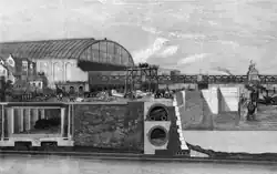 Illustrated section showing below ground structures of a riverside embankment including a covered railway line with steam train, riverside wall with sewer and pipes running behind. A large, glass roofed railway station sits in the left middle distance adjoining a railway bridge that crosses the river. Boats ply the water and tiny figures are engaged in construction work with a raised scaffold in the centre.