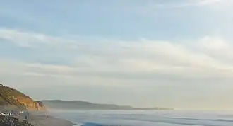 Finger of a mountain visible behind a cliff and on a beach with a partly cloudy sky