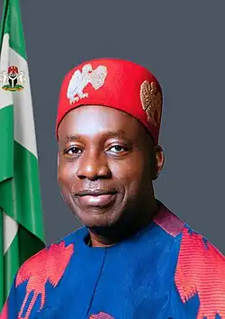 close-up image of Charles Soludo wearing a traditional colourful outfit and hat, smiling directly at camera, with Nigerian flag to the right