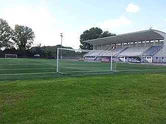 A football pitch with a goal net in the foreground, and a grandstand on the left